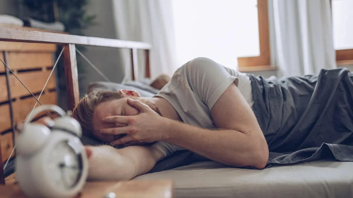 Man lying in bed the morning after drinking with his hand over his eyes, next to an alarm clock.
